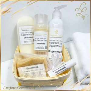 Luxious Lemon Unscented Self-Care Gift Set displayed in a small wooden basket. Includes five fragrance-free products: hand & body liquid wash, lotion soufflé, solid skin rescue salve, goat’s milk bar soap in an organza bag, and lip butter. Presented on a white surface with a folded towel in the background, emphasizing a clean and gentle skincare routine.