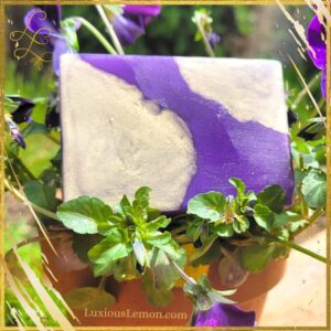 A luxurious handcrafted glycerin bar soap in deep purple and shimmering gold hues, nestled among vibrant green leaves and delicate purple flowers. The soap, named Take Ten Lavender is featured with a golden Luxious Lemon border, highlighting its artisanal beauty.