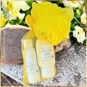 Yellow mesh gift bag containing two travel-sized bottles of Lemon Pound Cake scented hand and body wash and lotion soufflé, with a matching yellow shower pouf, displayed on a rock with white flowers in the background.