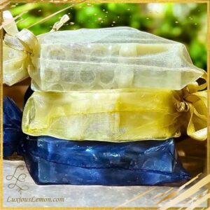A stack of three elegantly packaged bar soaps in sheer organza gift bags. The top soap is wrapped in a delicate ivory-colored bag, the middle one in a golden yellow bag, and the bottom soap in a deep navy blue bag. Each bag is tied with a matching ribbon, adding a luxurious touch. The background features a softly blurred green outdoor setting.