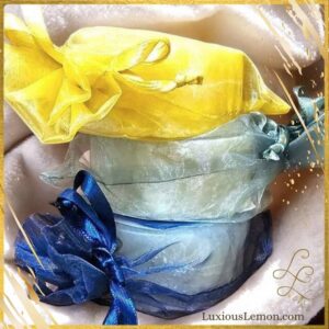 A stack of three Luxious Lemon Shave & Shampoo Bars, individually wrapped in sheer organza gift bags in navy blue, light blue, and yellow, tied with matching ribbon bows. The bars are stacked on top of each other and placed on a soft cream towel.