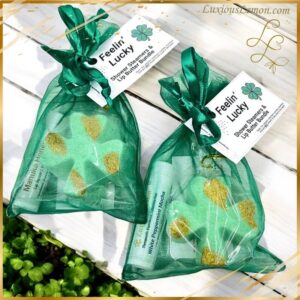 A St. Patrick’s Day-themed self-care bundle featuring a green organza bag filled with two clover-shaped shower steamers and a nourishing lip butter. The tag reads “Feelin’ Lucky” with a shamrock design. The bundle is placed on a white wooden surface with greenery in the background.