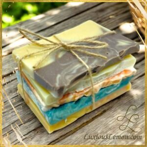 A rustic bundle of colorful handmade soap sample bars tied together with natural twine, resting on a wooden surface. The soaps are layered in various shades including yellow, brown, white, orange, and blue, with a marbled texture. The logo “LuxiousLemon.com” appears in the bottom right corner