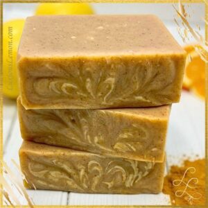 Stack of handcrafted Turmeric Tangerine Goat Milk Soap bars with a warm golden-orange hue and subtle swirl design, surrounded by fresh lemons, tangerines, and a sprinkle of turmeric powder in the background. The soap has a creamy texture and a natural, sunny glow that reflects its citrus and turmeric ingredients.