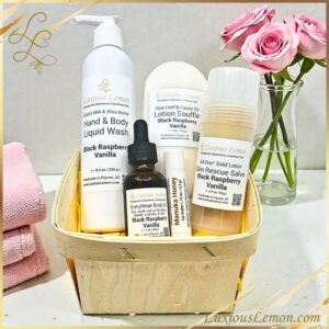 Natural self-care gift set in a small wooden basket featuring five products from Luxious Lemon, including a hand & body wash, lotion soufflé, solid salve, body oil dropper bottle, and lip butter. Displayed on a white counter with a vase of roses and pink towels in the background.