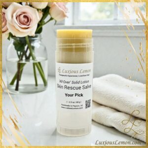 Luxious Lemon Skin Rescue Salve in a clear twist-up tube, labeled “All-Over Solid Lotion – Your Pick,” placed on a white bathroom counter with a vase of roses and rolled spa towels in the background.
